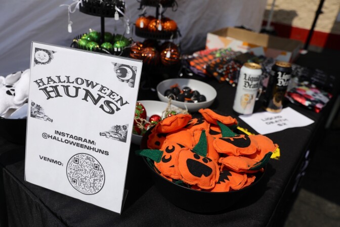 A table with a black tablecloth and a sign reading "HALLOWEEN HUNS; INSTAGRAM: @HALLOWEENHUNS; VENMO:" followed by a QR code. Decorations for sale include cloth pumpkins, green and orange Christmas ornaments, fabric ghosts, and more.