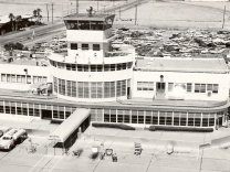 A black and white photo shows a white building flanked with bay windows and a tower on top. Cars are seen in the distance in a parking lot with the tale of an airplane seen in the lower left corner. 
