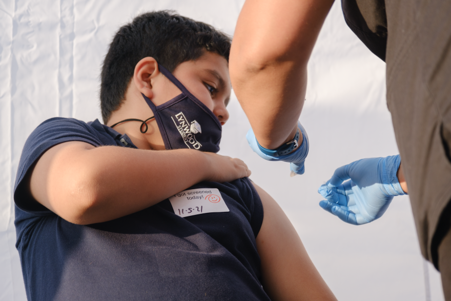 A boy in a navy blue shirt holds up his sleeve while his arm is cleaned in preparation for a coronavirus vaccine.