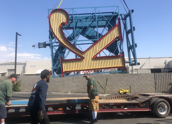 Three workers surround a flatbed truck, as a gigantic sign of the letter "K" -- tilted on its side -- is being lowered onto it