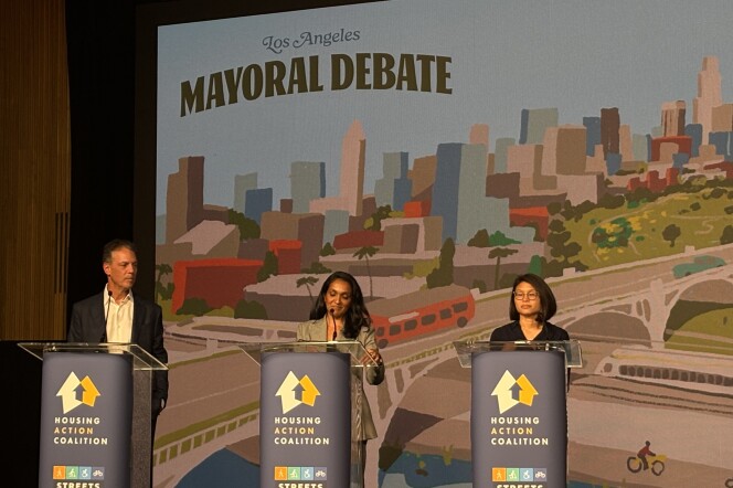 Three people stand at podiums on a stage. The podiums are branded with the hosts of the L.A. Mayoral debate held on March 23, including Housing Action Coalition and Streets for All. The background is an illustration of Los Angeles. It shows an orange bus on an elevated road with buildings, colored in red, brown, grey and blue, in the background. Below the elevated road is a biker cycling alongside water and a white train on the other side of the path.