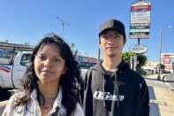 A male presenting person, wearing a black cap, and a female presenting person stand on a sidewalk.