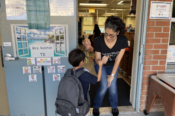 A woman in a black shirt and medium-tone skin pinky shakes a boy wearing a large gray backpack. A door shows different cartoon images of greetings like a hug, wave and fist pump. 