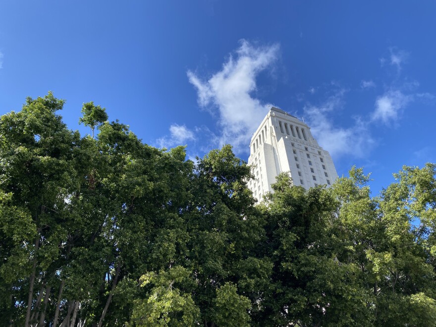 the top of LA City Hall can be seen against a blue sky and clouds with green leafy trees in the foreground. 