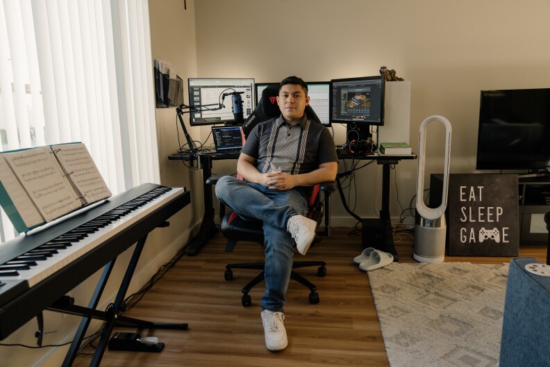 A Filipino man wears a short sleeve black and gray shirt, jeans, and white sneakers while sitting at a gaming chair. Behind him there's three computer screens, a lap top, and a microphone. To the left of frame there's a keyboard with sheet music. On the right of frame there's a TV and a print that reads "Eat, Sleep, Game."