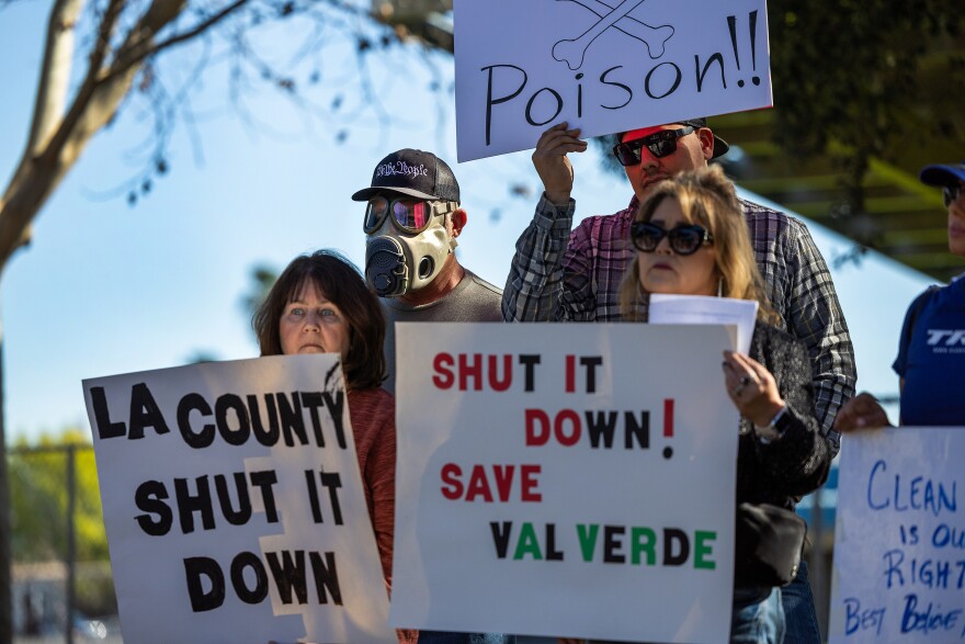 A group of people, including a woman on the left, young boy in the middle, and an adult man on the right, are holding signs up in the air during a protest. The young boy, who is wearing a bright blue t-shirt, is holding a sign that reads "shame on you! you smell like poo!" The adult man, who is wearing a gray t-shirt, is holding a white sign with black text that partially reads "shut it down."