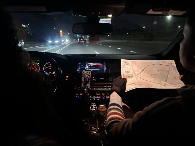 The interior of a car from the vantage point of the backseat. The person on the right in the passenger seat is holding up a printed map with both hands, which are wearing fingerless black gloves. The interior is slightly illuminated by buttons on the dashboard, with streetlights and car headlights shining through the front windshield. 