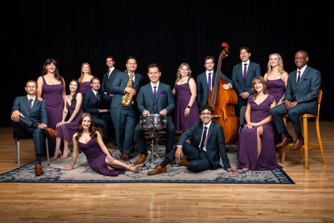 A group of 16 people people all posing. From left to right: A light-skinned man wears a navy blue suit and purple tie with brown shoes and sits on a chair with legs crossed. To the right, a light-skinned woman stands next to him wearing a purple dress. Another light-skinned woman wearing a purple dress is sitting next to her. A light-skinned woman is partially laying down on a blue and white rug also wearing a purple dress. To her right, a light-skinned man wearing a navy suit sits on a chair and places hands on a drum. To the right, a medium-light toned man sits on the rug wearing a navy suit. To his right, a light-skinned woman wears a purple dress and sits on a chair. To her right, a dark-skin toned man sits on top of a tall chair. To his left, a light-skinned woman wearing a purple dress. To her left, a light-skinned man wears a navy suit. To his left, a light-skinned man wearing a navy suit holds a cello. To his left, a light-skinned woman with blonde hair wears a purple dress. To her left, behind the man with the drum, a light-skinned man wearing a navy suit holds an alto-saxophone. To his back left, a medium-light skin toned man stands wearing a navy suit. In front, a light-skinned toned man wearing a navy suit sits on a chair. Behind him, a light-skin toned woman stands wearing a purple dress. 