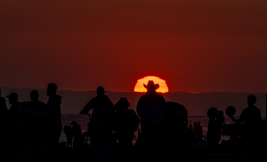 Large crowds are silhouetted by the setting sun as they celebrate Independence Day before watching fireworks displays shooting off a pier and illuminating the ocean.