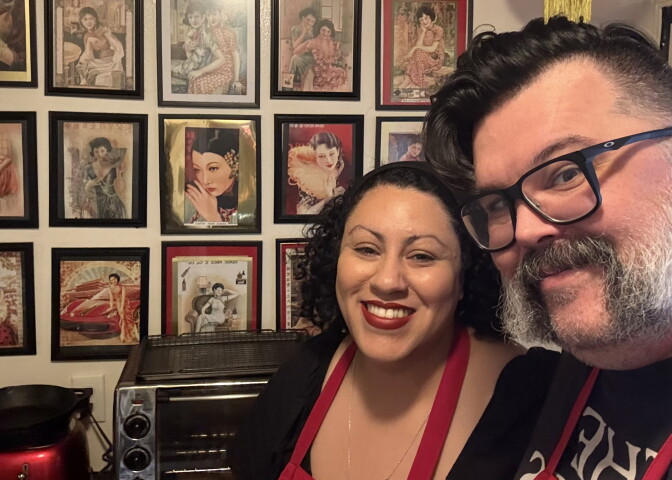 A woman with light brown skin and dark, curly hair, and a man with light skin, glasses, and a graying beard, both wearing red aprons, smile in a kitchen, standing in front of a wall lined with framed vintage illustrations.