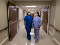 Two nurses, one in deep blue scrubs and the other wearing light blue, stand in a hospital corridor with masks on.