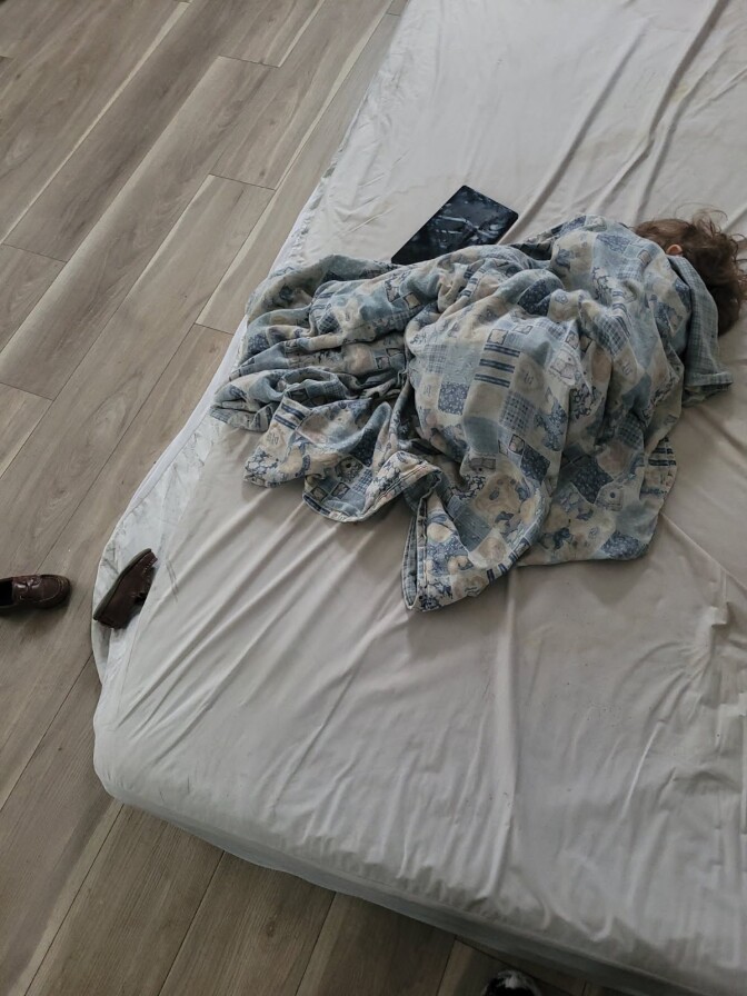 A young boy curled up under a blue and white blanket sleeps on a mattress on a wooden floor.