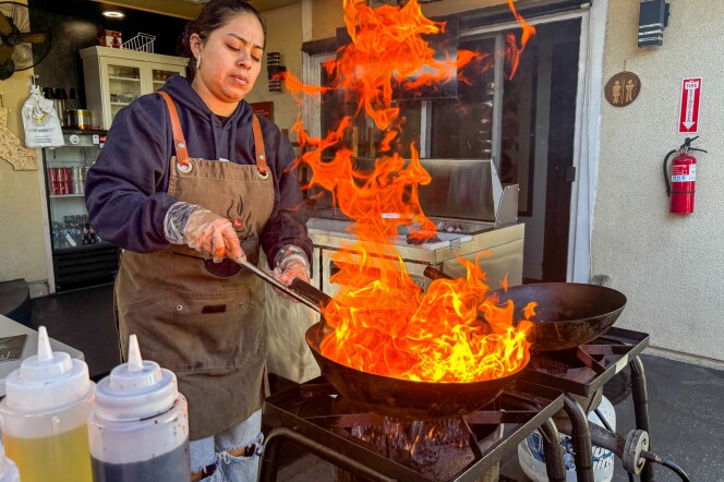 A woman in a brown Lomo Fuego apron stirs a wok over a powerful outdoor burner, producing dramatic flames that leap several feet into the air in a backyard restaurant's  patio area. 