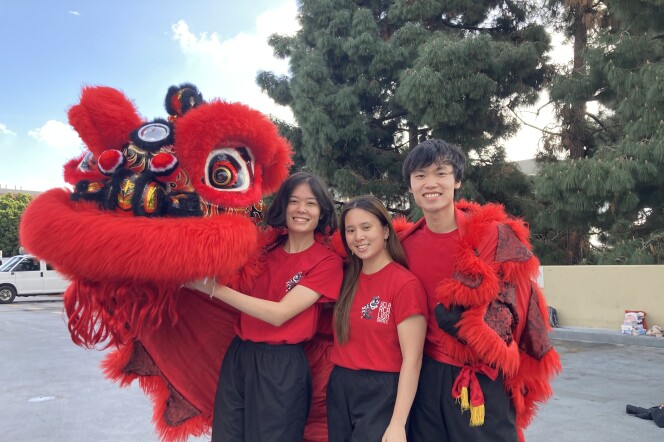 Three people with brown hair, red t-shirts and black pants pose holding a red, furry Chinese lion dance costume. They're standing in a parking lot with a car and a tree top in the background.