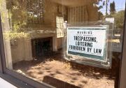 A front window in a home, with the bare living room and fireplace seen inside. A black and white sign that reads "Warning state property trespassing, loitering forbidden by law" is taped on the front facing out towards the street.