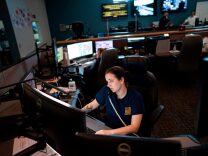 A woman with dark hair in a ponytail and wearing a blue t-shirt sits at a desk in front of several computer screens answering 911 calls.