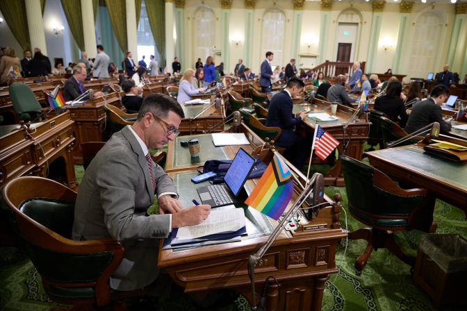 A lawmaker, wearing a gray suit and glasses, sits at their desk on the Assembly floor marking a sheet of paper with a pen.