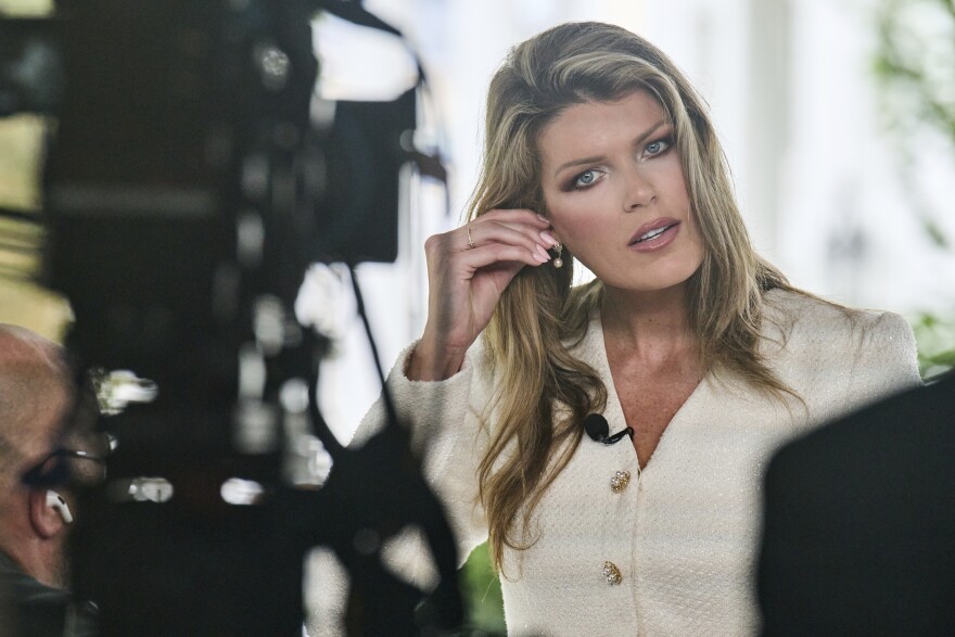 Lindsey Halligan, a woman with light skin tone and blonde hair, wearing a white coat, puts in an earing as she stands in front of a camera pointed at her, which is out of focus in the foreground on the left.