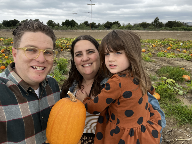 A man wearing a plaid shirt and glasses holds a pumpkin next to a woman with long brown hair, holding a little girl with brown hair.