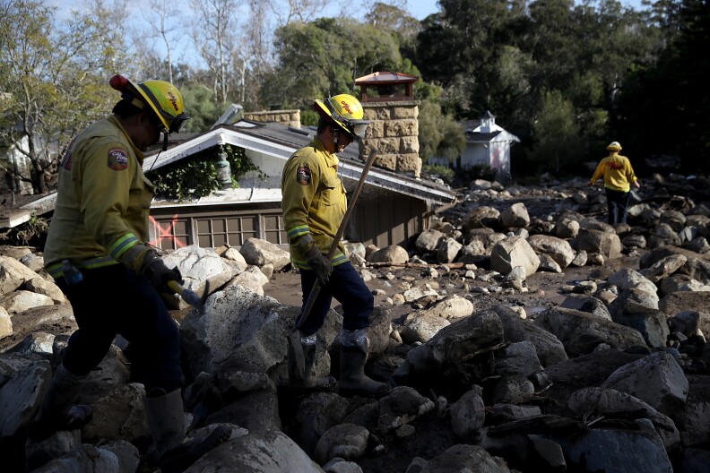Firefighters search for people trapped in mudslide debris on top of rocks and mud with a partially buried home in the background.
