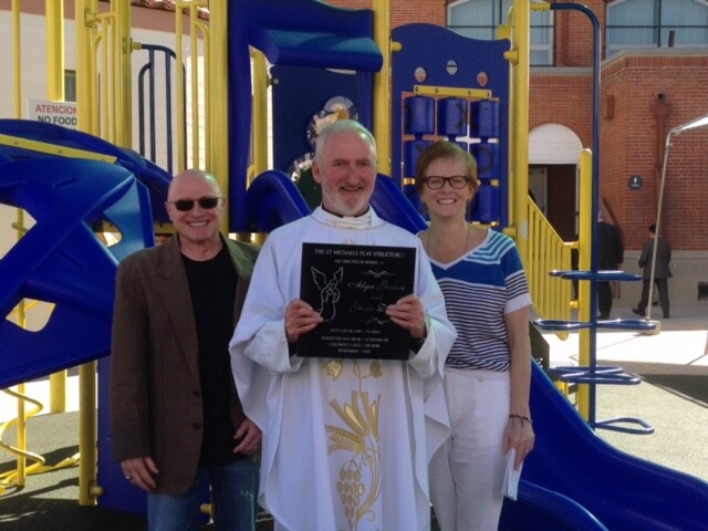 A man in a white robe holds a plaque. Two people stand on either side in front of a play structure.