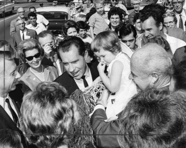 A black and white view of Nixon, a white man, carrying a child as he moves through a crowd of people surrounding him. There are mostly people with light skin tones by him, smiling and taking photos.