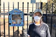 A woman wearing a muted green baseball cap, a white face mask, and black t-shirt is looking ahead into the camera. She's standing next to a blue, purple, and pink wooden box affixed to a black metal gate in front of a several story tan building.