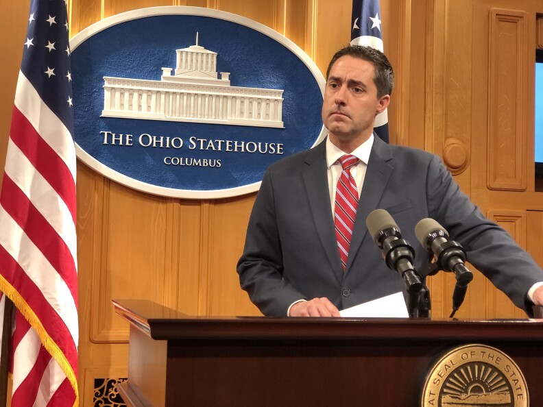 A white man in a red tie and dark suit stands at a lectern in front of a wall sign that reads: The Ohio Statehouse Columbus