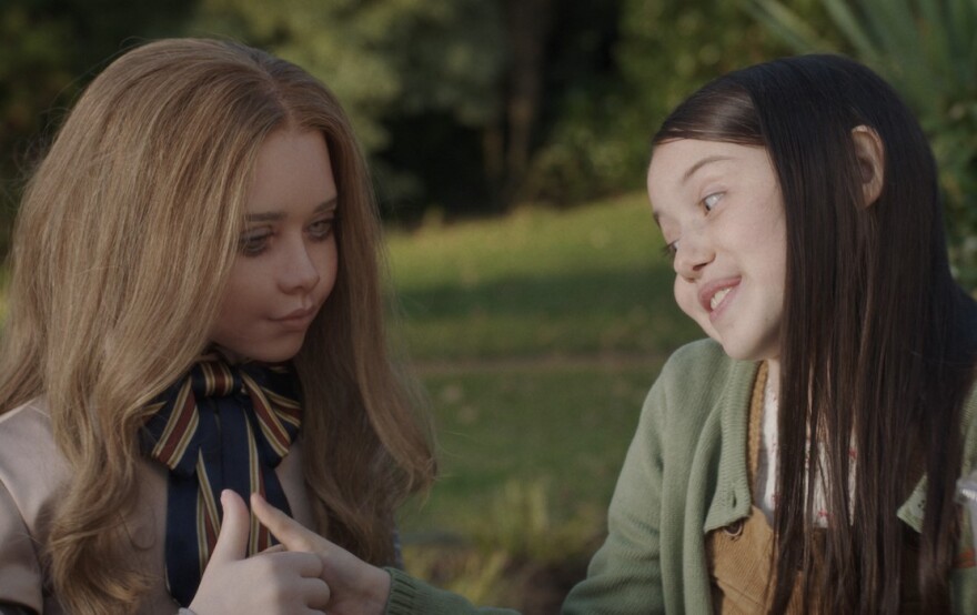 Amie Donald and Violet McGraw sit together outside with green grass and trees behind them. Donald sits on the left wearing a tan tailored dress with a large bow tie with blue, red, and gold stripes. The tails of the bow tie are long, falling down her shirt front. Her long blonde hair falls loose at her shoulders. McGraw wears a light green cardigan with tan overalls peaking through over a white t-shirt. Her long brown hair creates a straight curtain across the front of her left side. The two girls hold hands, with each of their thumbs raised, seeming to be playing a game of thumb war. Donald stares blankly past McGraw, her appearance robotic and artificial. McGraw focuses intently on their interlocked hands, her eyes are wide and a toothful smile across her face.