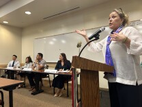 A woman stands at a podium and speaks to the ground, while four individuals — all candidates — sit at a nearby table at the front of a crowded room. 