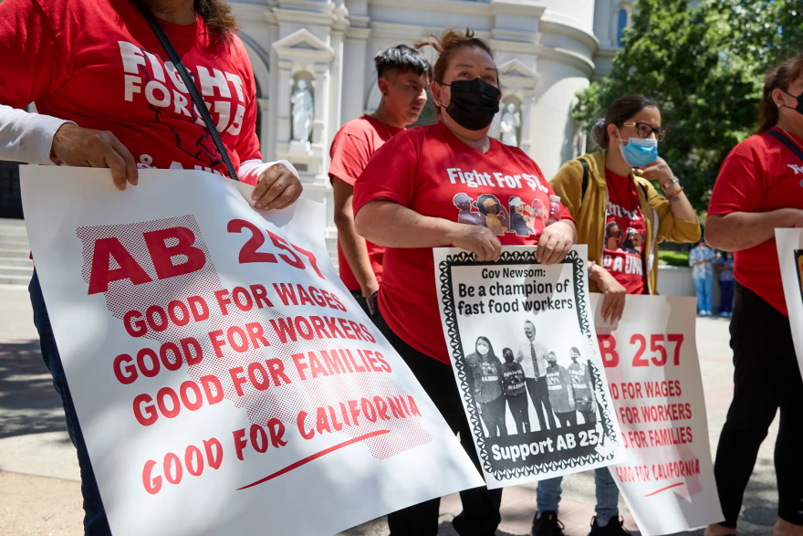 Protesters stand outside wearing red shirts and masks and carry signs in favor of AB257.