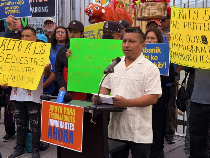 A man with medium skin tone and short black hair wears a an embroidered, white collared shirt. He is standing behind a podium with a microphone, holding a sheet of paper in his hands. Behind him, about a dozen people hold up signs that read: "No more raids," "No more family separations," and the like. 