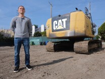 A man with light-tone skin stands with his hands behind his back in a dirt lot. A CAT bulldozer is behind him. 