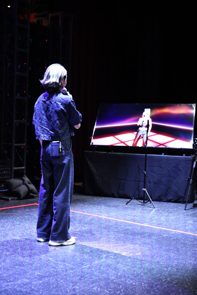 A young woman with light color hair dressed in denim holding a mic. She is standing and performing in front of a screen displaying a virtual avatar.