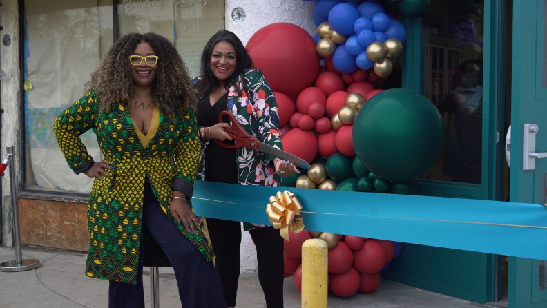 two women stand in front of a store next to a large teal ribbon. one of them holds an oversize pair of scissors.