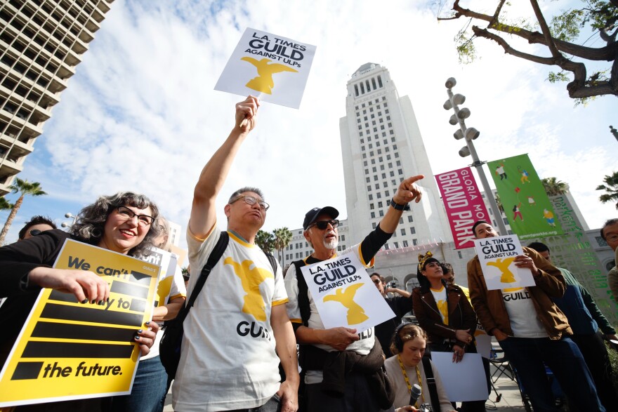 A group of people, some wearing white shirts with a yellow eagle and black text that reads "Guild" gather outdoors holding white, yellow, and black signs that reads "L.A. Times Guild Against ULPs." Los Angeles City Hall is in the background.