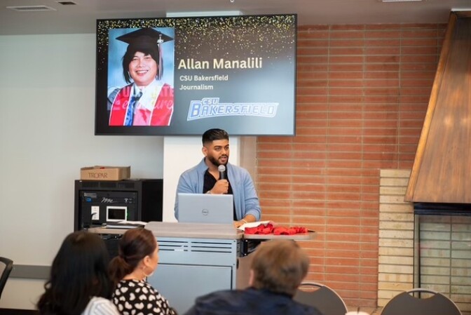 A man with medium brown skin, short dark hair, and a beard stands at a lectern in front of a classroom. He's holding a microphone with his laptop open in front of him. Behind him is a screen with a picture of a smiling person with a graduation cap and the words "CSU Bakersfield."