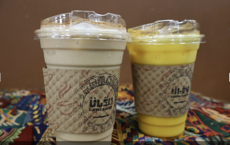 Two cold drinks stand side by side on bright colored fabric. On the left is a plastic cup containing a light brown liquid, and on the right is a plastic cup with a bright yellow liquid. Both have paper holders on the outside that say Ulew, surrounded by ancient Mayan symbols