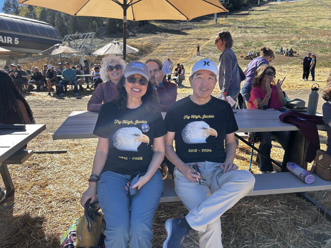 Two people wear black T-shirts reading 'Fly High, Jackie, 2012-2026' with an eagle image, sitting at a picnic table near a ski lift.