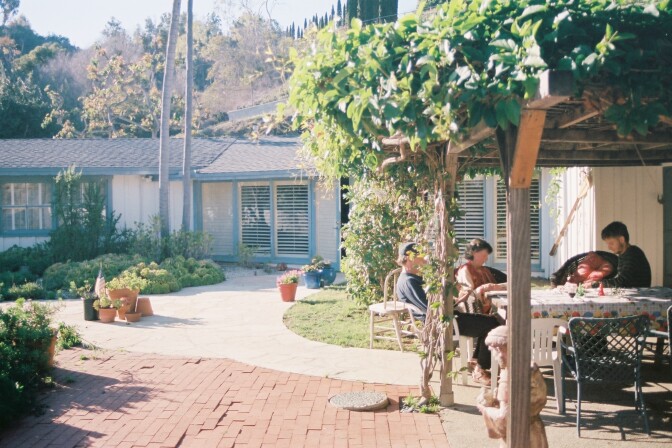A family sits outside under an awning covered in green vines. It's a sunny day. A house and palm trees are in the background.