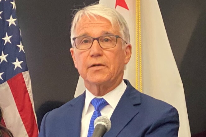 George Gascón, a light-skinned Latino man with white hear and wearing glasses, speaks in front of a microphone. He's wearing a blue suit jacket, blue tie, and white shirt.