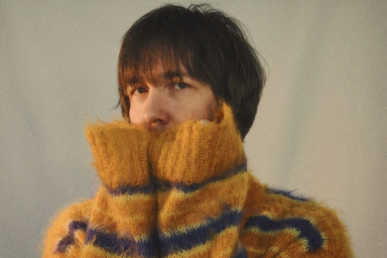 Aaron Lee Tasjan wears an orange, blue, and yellow striped wool sweater. He's covering half of his face with his sleeves, so the view sees just his nose, eyes, and brown bowl haircut.