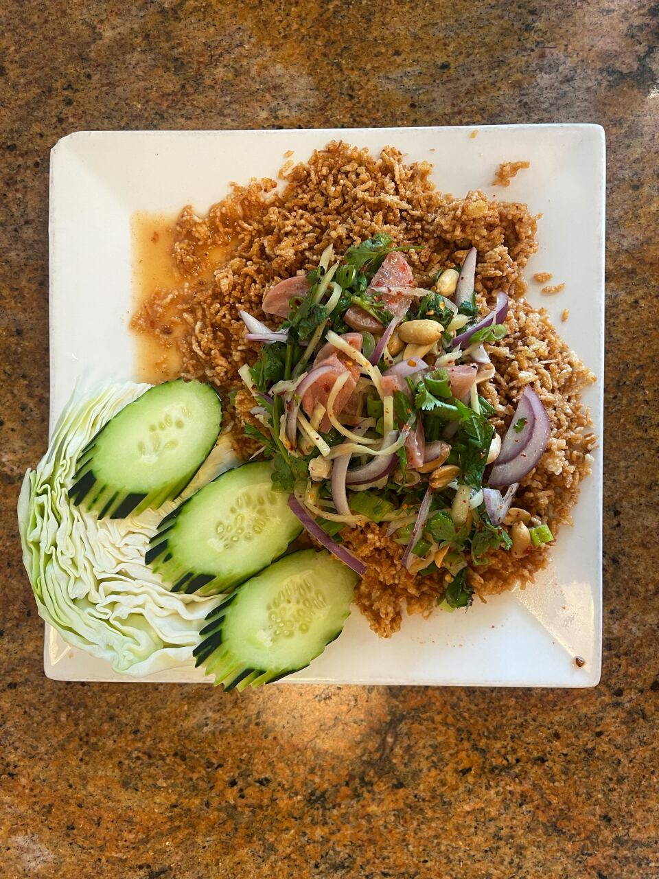 A top view of a white square plate with a pile of rice and red onions, herbs and peanuts. Next to it are three slices of cucumber and some cabbage