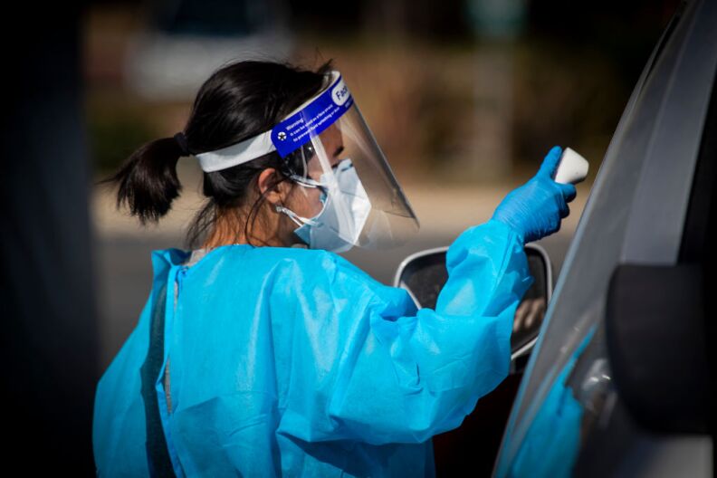 A woman with a short dark ponytail who is wearing a plastic face shield, plastic blue gloves and medical gown holds a digital thermometer up to a car window.
