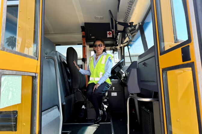 A woman with medium skin tone wears a bus operator uniform, including a neon green vest. She stands up against the driver's seat for a portrait taken through the open doors of a yellow school bus.