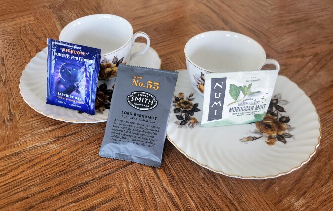 Two porcelain cups and saucers sit on a wooden table. Leaning on them are three tea bag packets which say Bigelow butterly pea flower, No. 55 Steven Smith teamaker and Numi Moroccan mint.