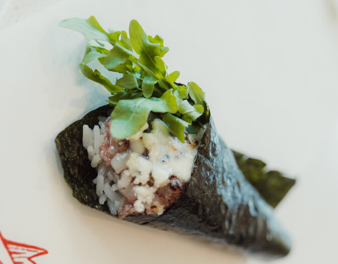 A hand roll wrapped in nori, filled with rice, dry-aged beef, melted cheese with visible char marks, and topped with a bundle of fresh arugula, photographed close-up against a white background.