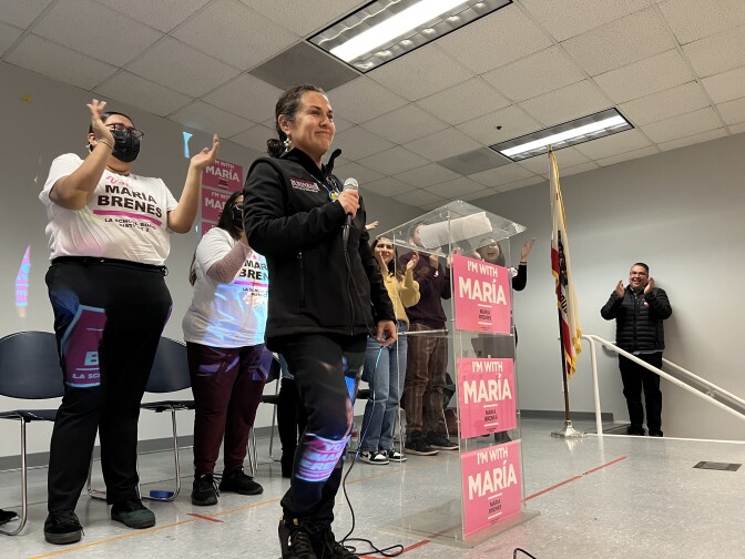 A brown-skinned woman wearing a black, light zip-up jacket and black pants holds a microphone and stands next to a podium with pink signs reading "I'm With María."