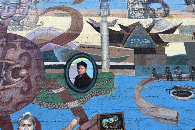 A section of an outdoor mural depicting Filipino American heritage. At the center of the photos is an oval portrait of a person with short hair, wearing a dark jacket, framed in green and blue. Around it are various scenes and symbols.