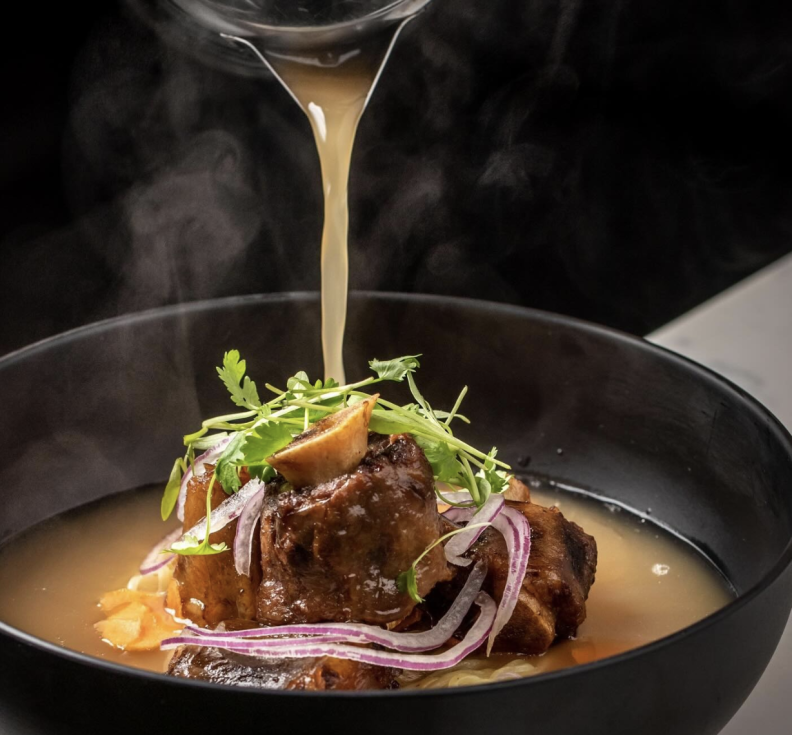 An attractive black bowl contains a deep rich brown broth. In the center is an oxtail, red onions and green herbs. Above it a jug is pouring the broth on top of the oxtail.